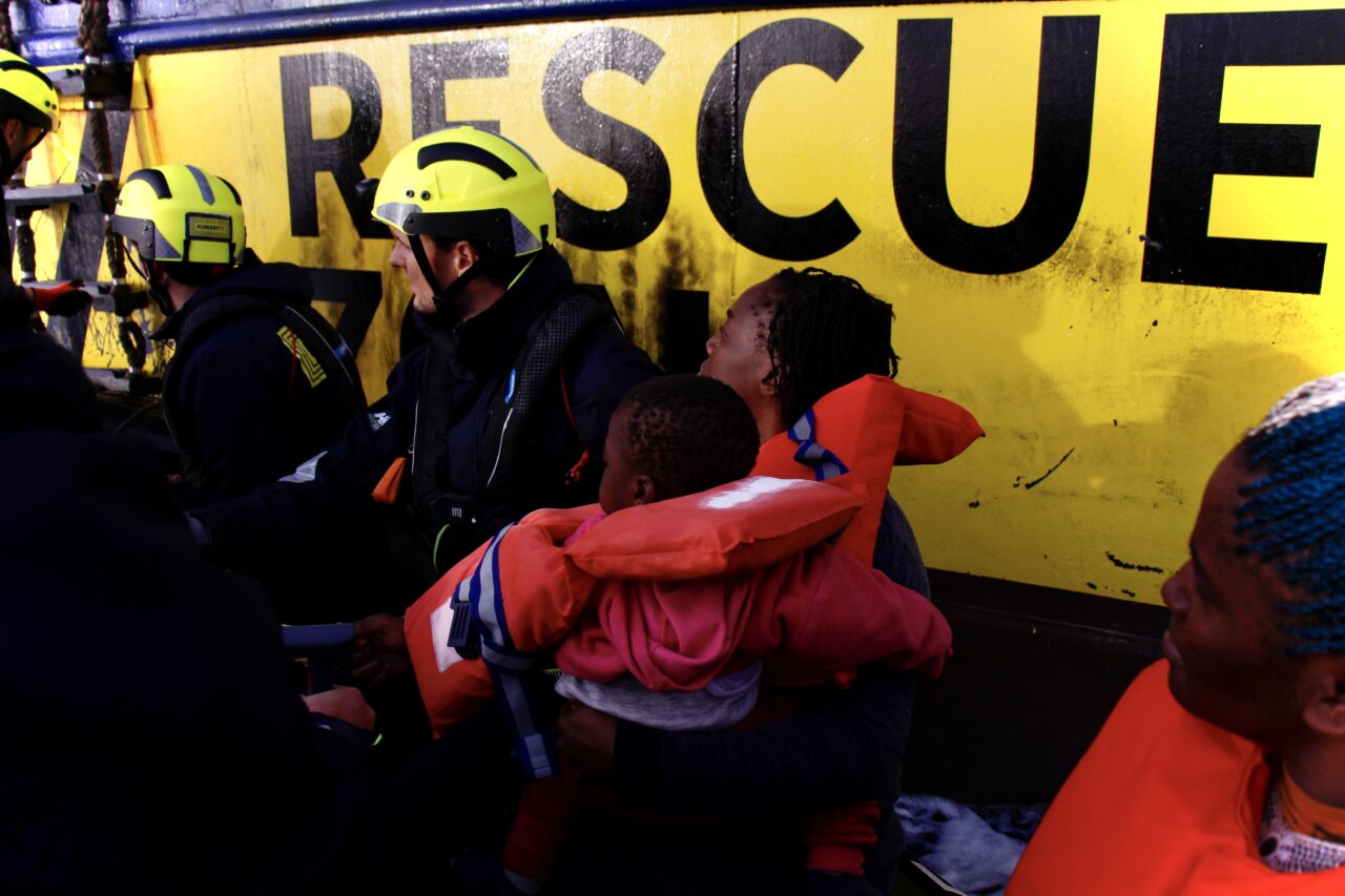 Vor der Bordwand der Humanity 1 sind links Frauen und Kinder in Rettungswesten, rechts Mitglieder des Rettungsteams zu sehen.