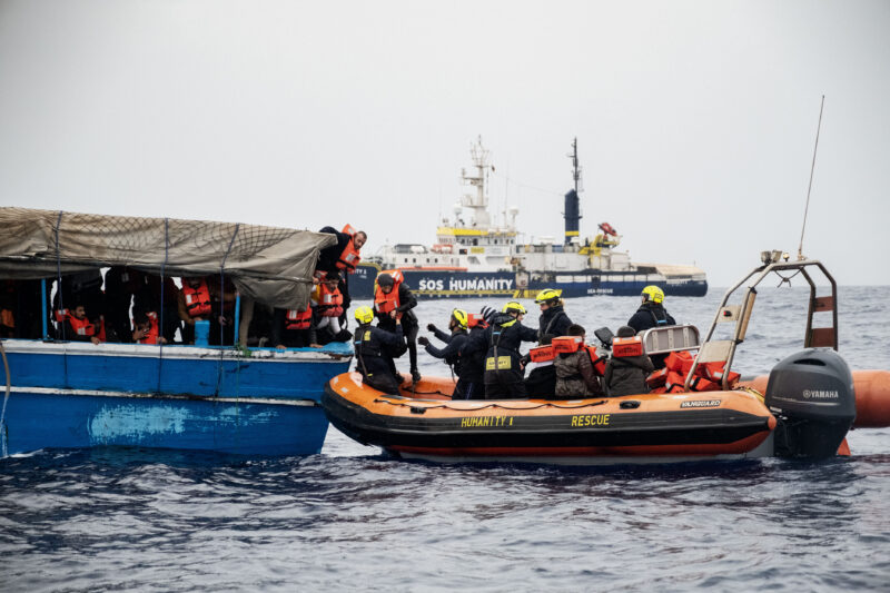 Geflüchtete Personen bei der Rettung von einem blauen Holzboot auf das RHIB der Humanity 1, das Rettungsboot von SOS Humanity.
