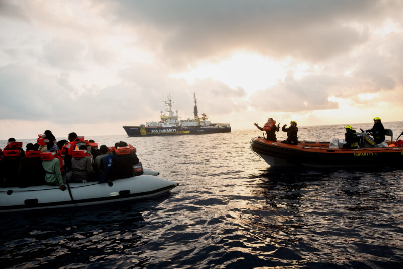 RHIB von SOS Humanity mit Crew, die Menschen von einem Schlauchboot rettet. Im Hintergrund das Meer, Himmel und die Humanity 1.