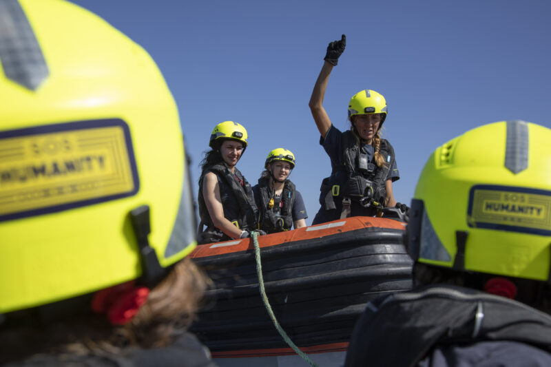 Drei weibliche Crewmitglieder von SOS Humanity auf einem RHIB. Im Vordergrund die Köpfe mit gelben Helmen von anderen Personen.