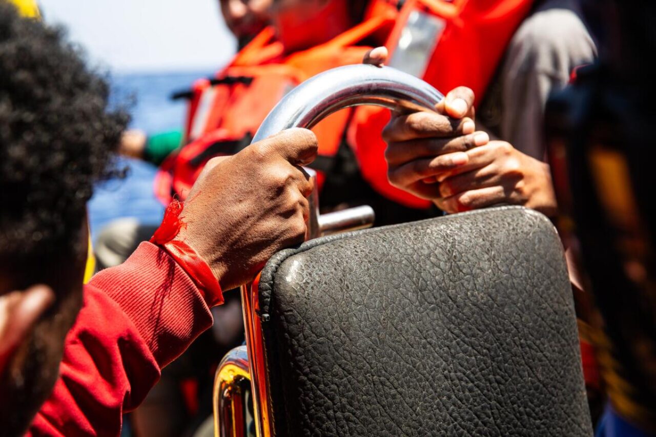 Zwei Hände, die sich an einem Griff auf dem Rettungsboot festhalten. Im Hintergrund sind Menschen in Rettungswesten.