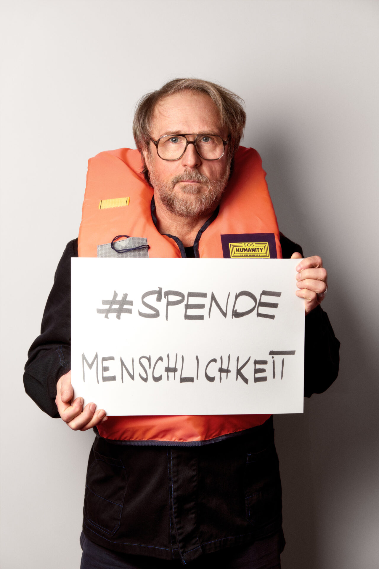 Bjarne Mädel mit einer Rettungsweste und dem Schild 'SPENDEMENSCHLICHKEIT